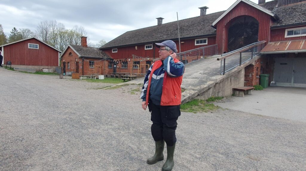 Maataloustyöhön sopivaan asuun pukeutunut nainen vanhan navetan edessä.