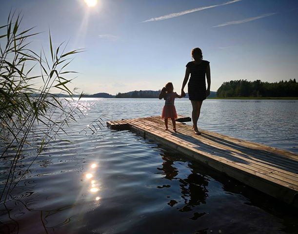 Kuvassa äiti ja lapsi kävelevät käsikädessä laiturillla, takana paistaa aurinko, on kesä.