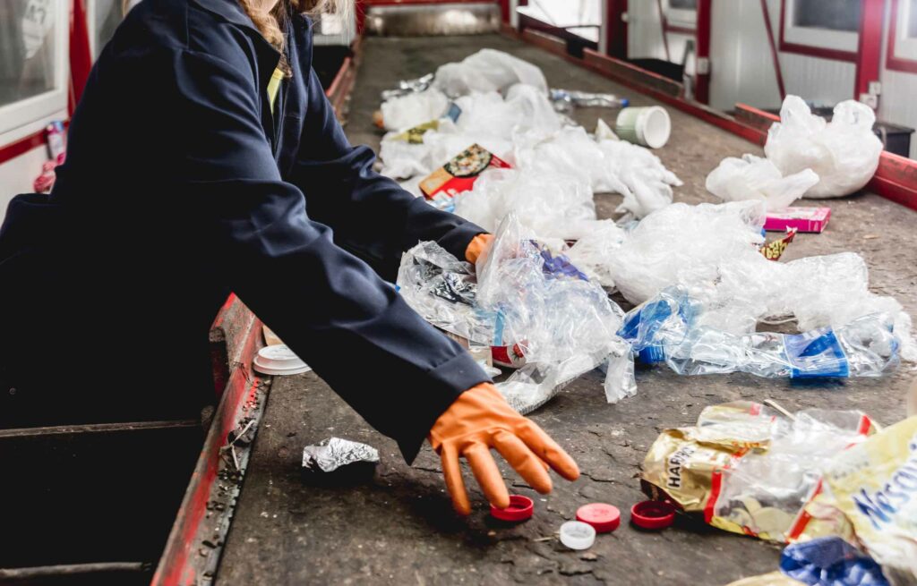 Kuva jätelaitokselta, jossa tummiin haalareihin ja oransseihin suojakäsineisiin pukeutunut työntekijä poimii liukuhihnan ääressä muoviroskien seasta sinne kuulumattomia jätteitä.