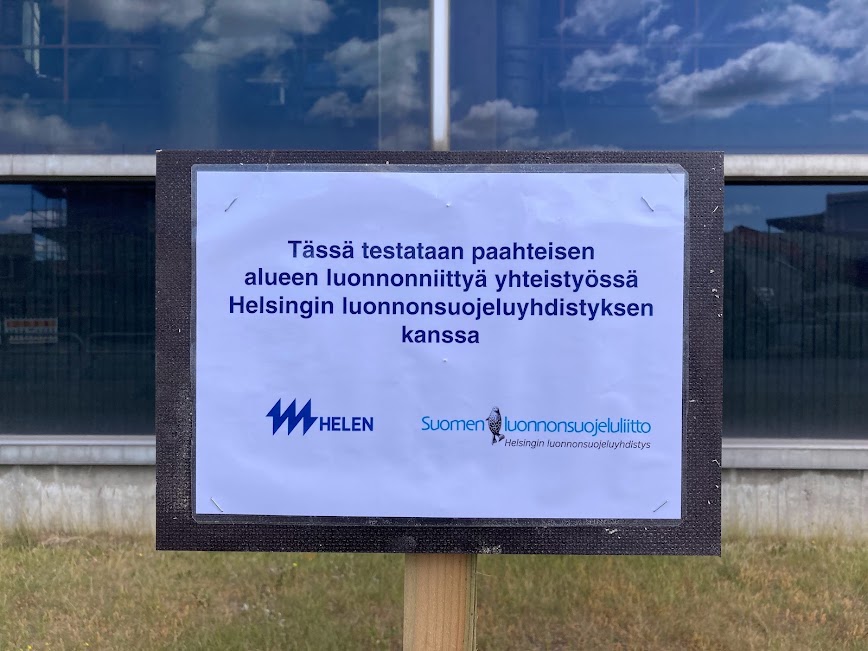 Kyltti, jossa teksti "Tässä testataan paahteisen alueen luonnonniittyä yhteistyössä Helsingin luonnonsuojeluyhdistyksen kanssa" sekä Helenin että SLL Helsyn logo.