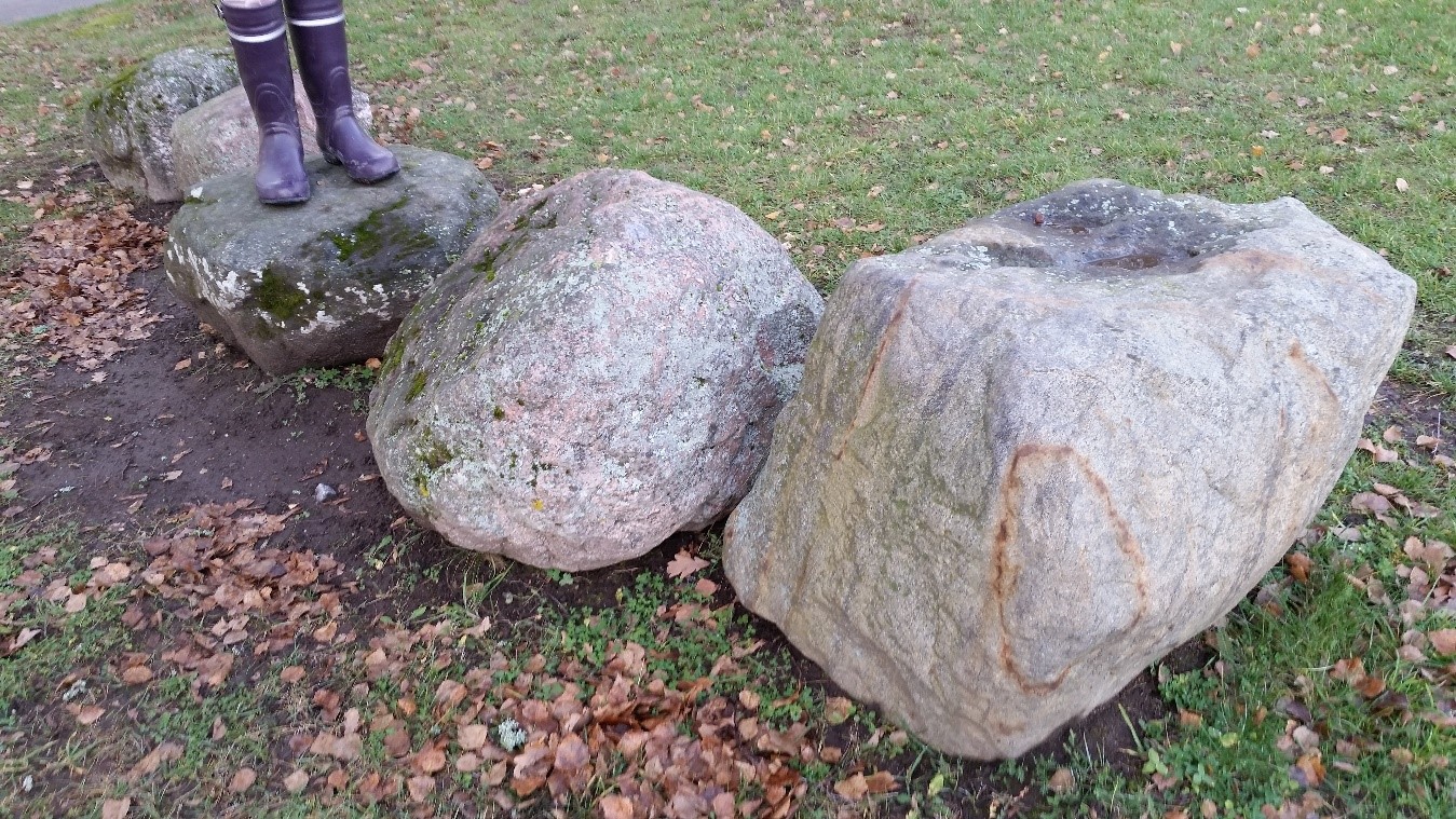 Vierekkäisiä kivenlohkareita nurmikolla. Yhden kiven päällä näkyy, jonkun tasapainoilijan violetit saappaat. Etualan kivessä erottuu ruskea, hapettuneesta rautapitoisesta mineraalista kertova raita.