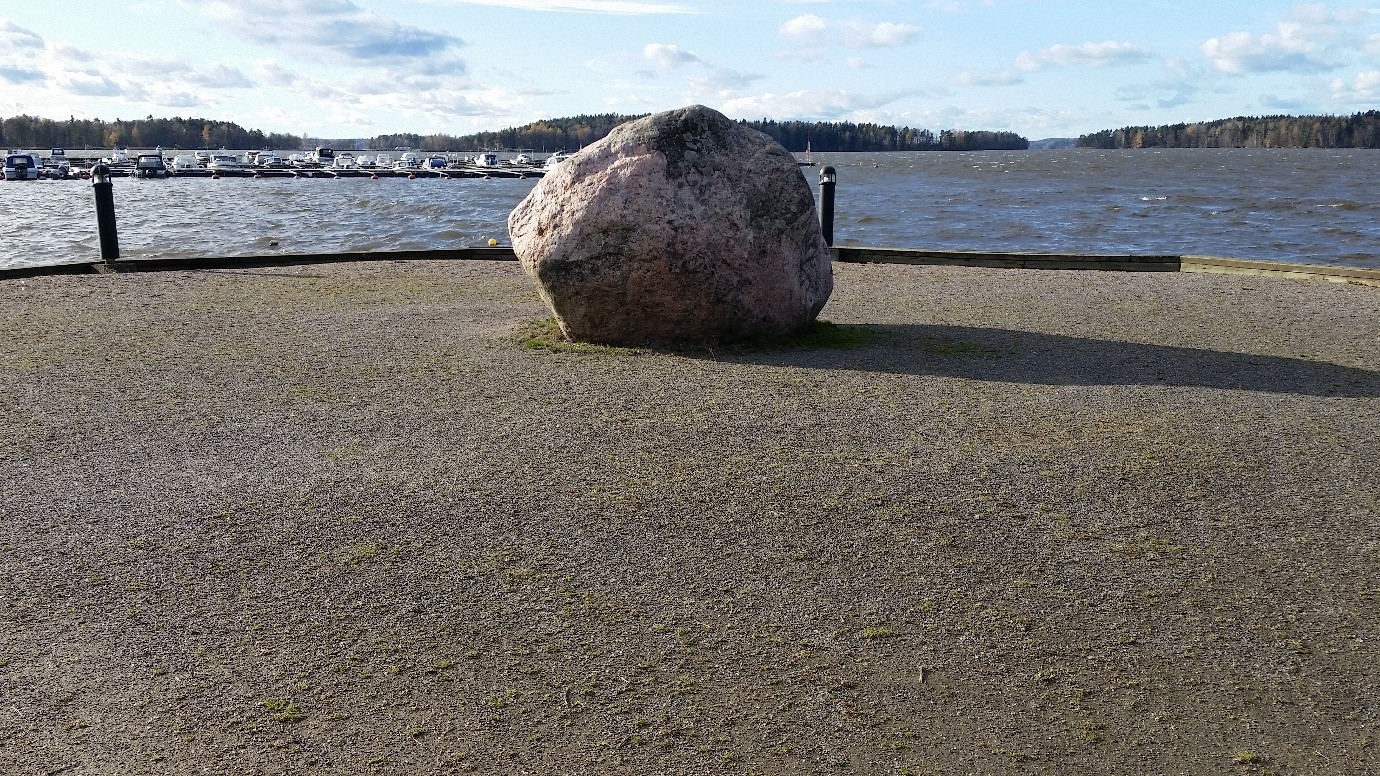 Niemen kärjessä on iso pyöreähkö kivi, joka koostuu graniittisesta pegmatiitista. Taustalla lainehtii Lohjanjärvi ja näkyy veneitä sataman laiturissa. Horisontissa näkyy puustoisia saaria. Sinisellä taivaalla on pieniä pilviä. 
