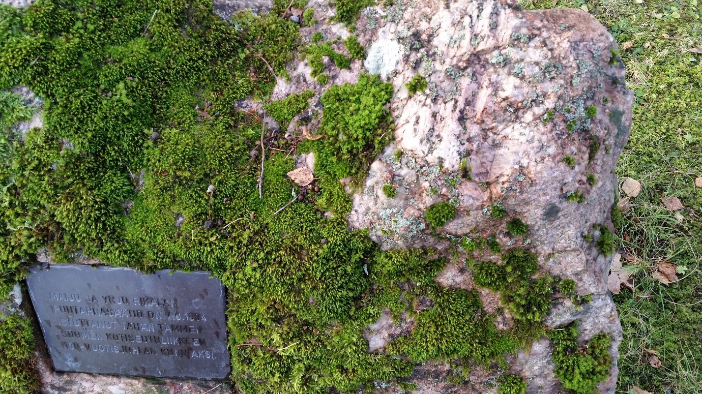 Punertavaa kivenlohkaretta peittää syvän vihreät sammalet. Sammalten keskeltä erottuu tumma muistolaatta. 