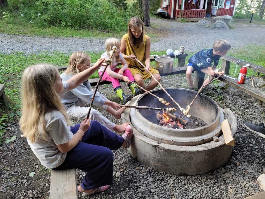 Leirinohjaaja ja nelj&auml; lasta paistavat tikkupullaa nuotiolla kes&auml;illassa.