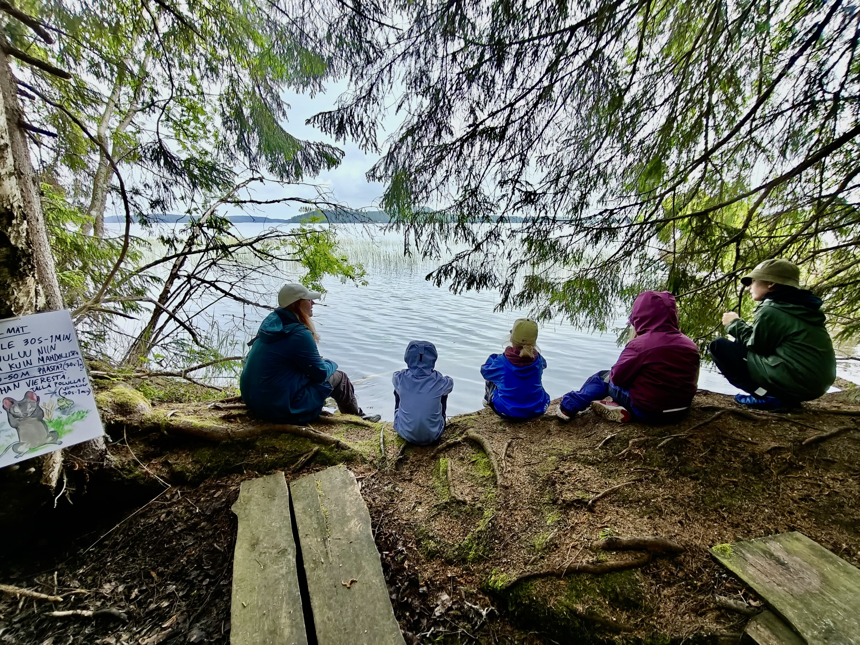 Ohjaaja ja neljä lasta istuvat järven rannalla puiden katveessa.