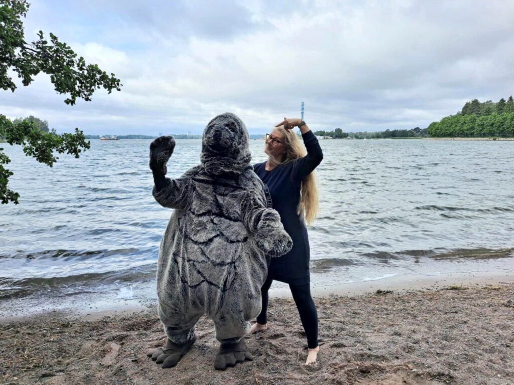Norppa-asuinen ihminen ja vaalea nainen hiekkarannalla. Taustalla ItÀmeri.