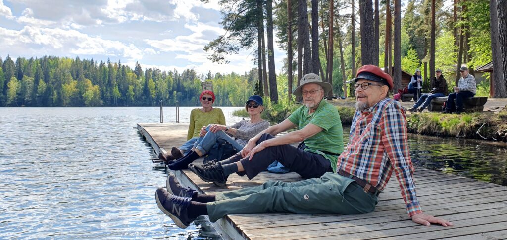 Helsyn retkeläisiä hengaa laiturilla Suujärven rannalla. Aurinko paistaa.