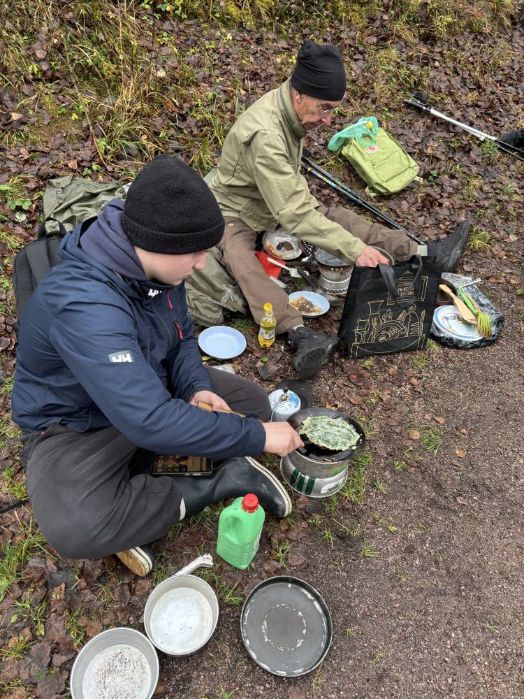 Två män sitter på marken i skogen och steker plättar på stormkök.