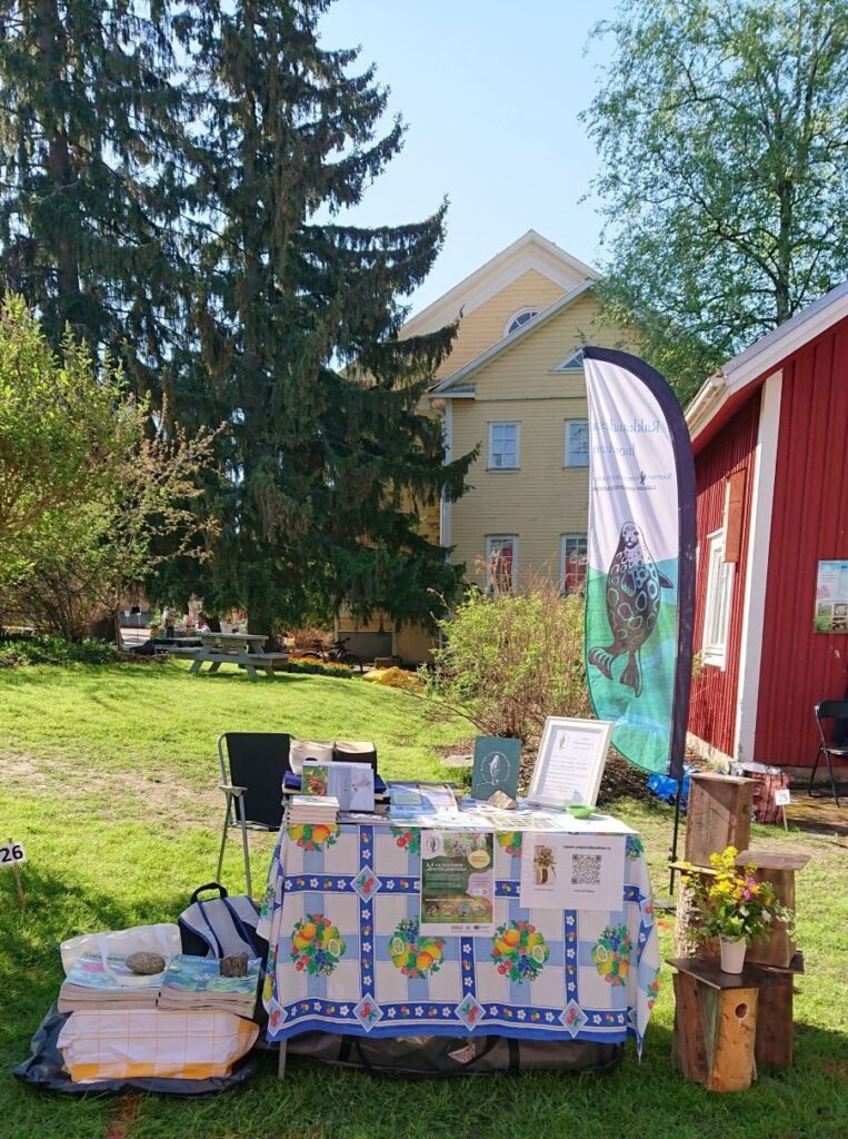 Messupöytä puutarhassa. Pöydän vieressä linnunpönttöjä ja takana yhdistyksen lippu. Taustalla suuria kuusia, koivu sekä keltainen puurakennuksen pääty.