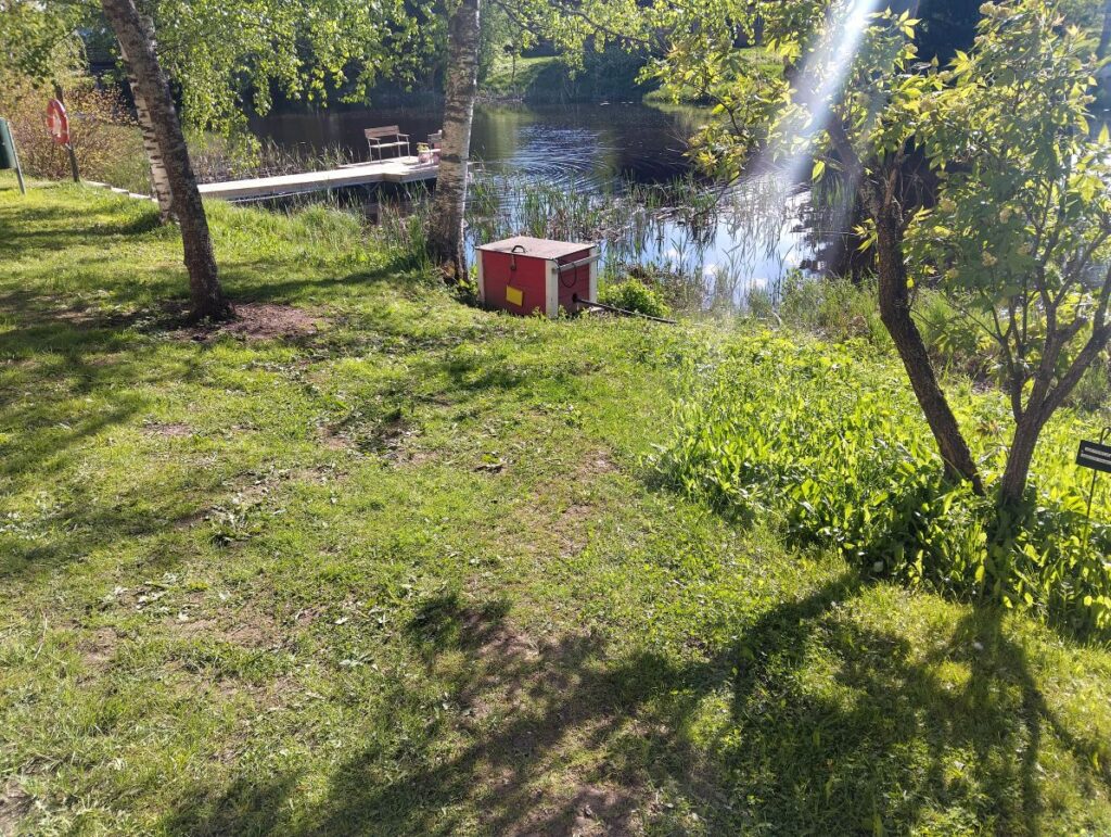 Joen rannassa koivun alla pieni punaiseksi maalattu laatikko nurmialueen reunalla. Laatikon kyljessä roikkuu keltainen kuvakortti niin, että kuva on kääntynyt laatikkoa vasten.
