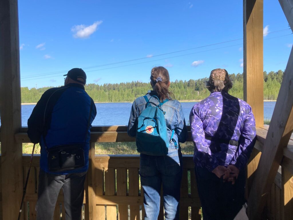 Kolme ihmistä katsomassa järvimaisemaa aurinkoisessa säässä