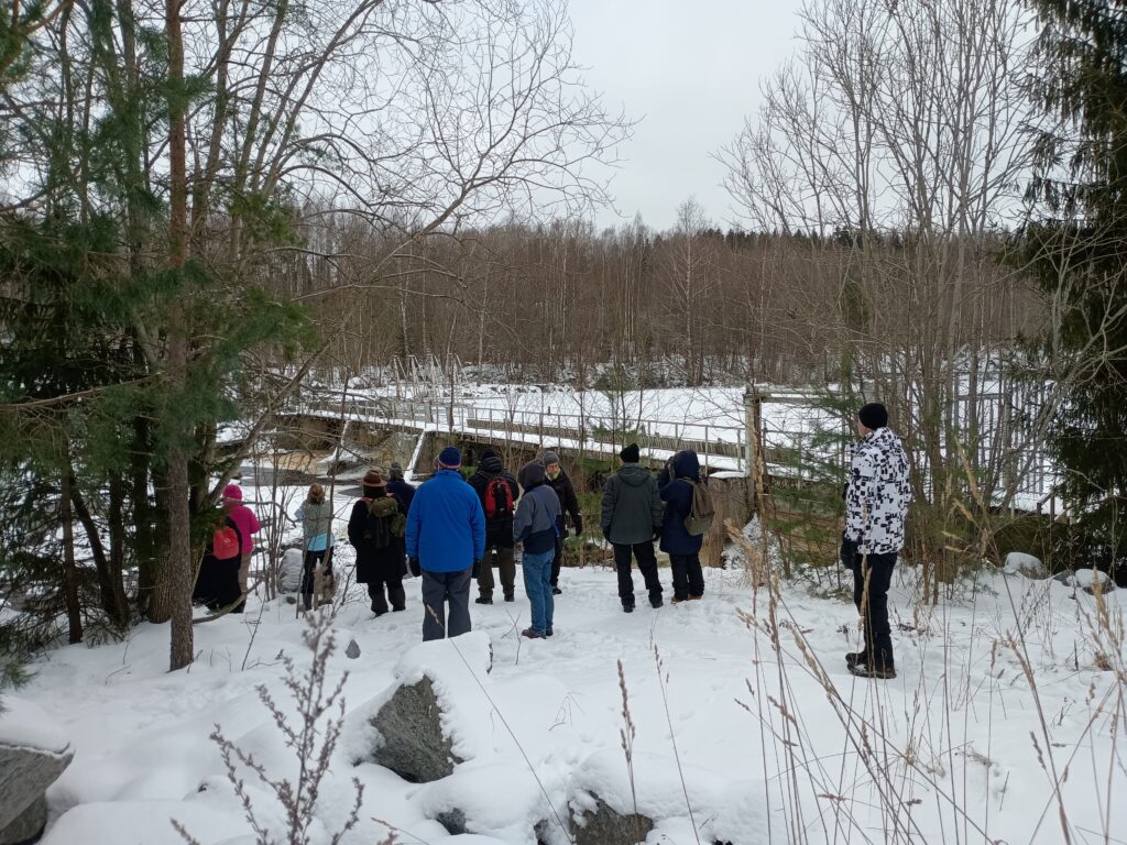 Ryhm&auml; ihmisi&auml; lumisella mets&auml;polulla tarkastelemassa jokea ja patoaluetta talvella. Opastettu retki.
