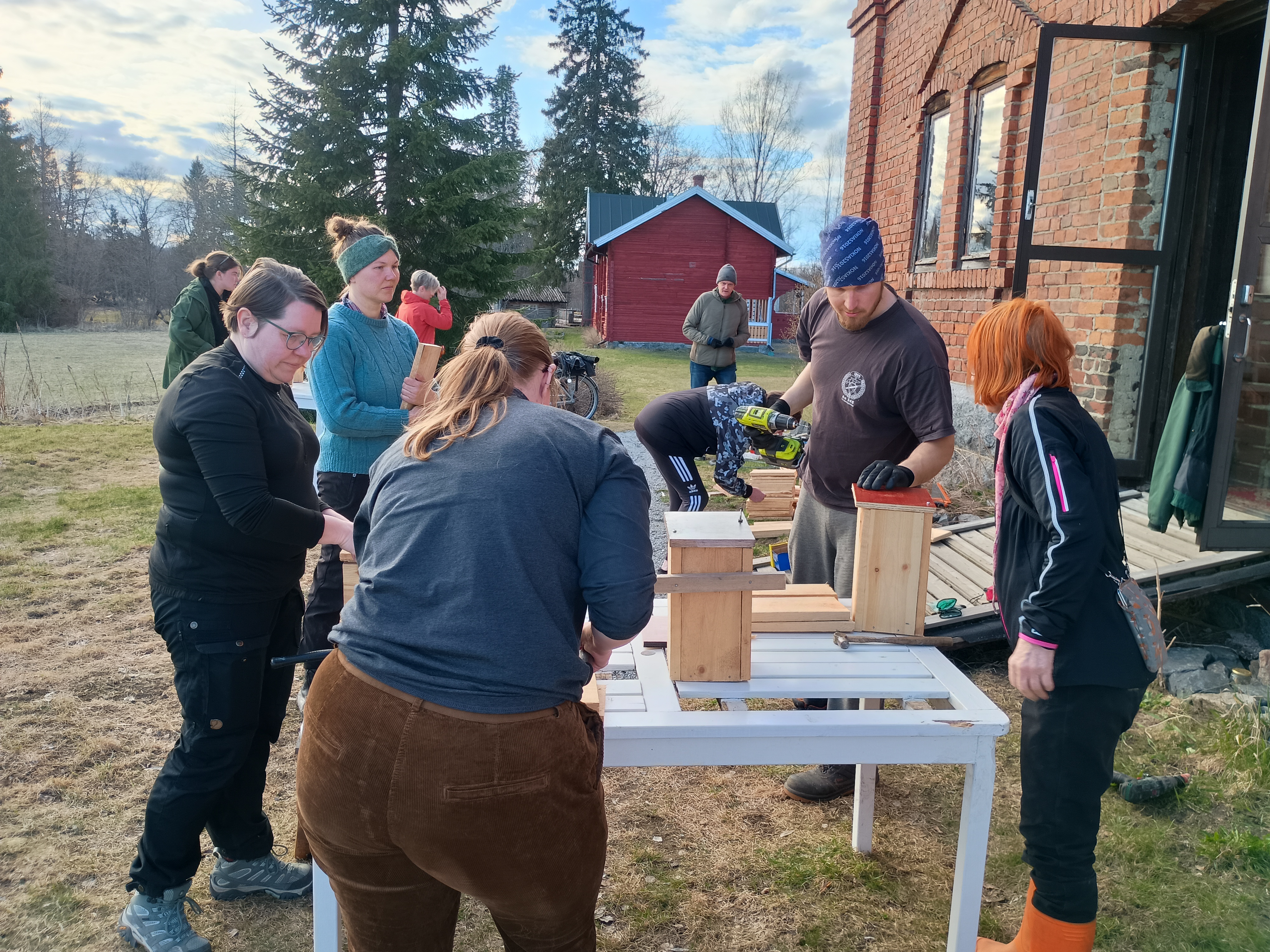 Ryhmä aikuisia rakentaa linnunpönttöjä ulkona valkoisella pöydällä. Yksi henkilö poraa akkuporalla, muut seuraavat ja avustavat. Taustalla näkyy punainen tiilitalo ja punainen ulkorakennus.