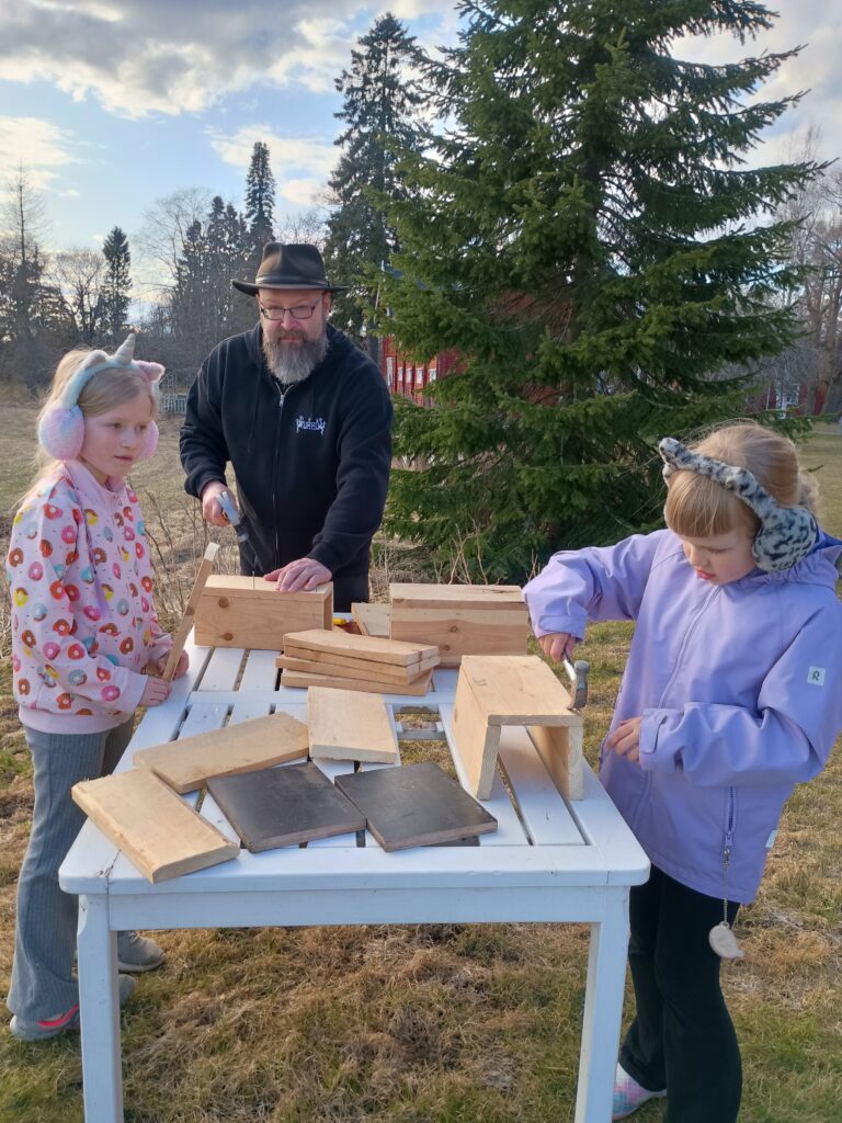 Parrakas mies mustan hatun kanssa ja kaksi nuorta tyttöä rakentavat yhdessä linnunpönttöjä valkoisella ulkopöydällä. Tytöillä on korvalaput päässä. Taustalla näkyy havupuita ja punainen rakennus.