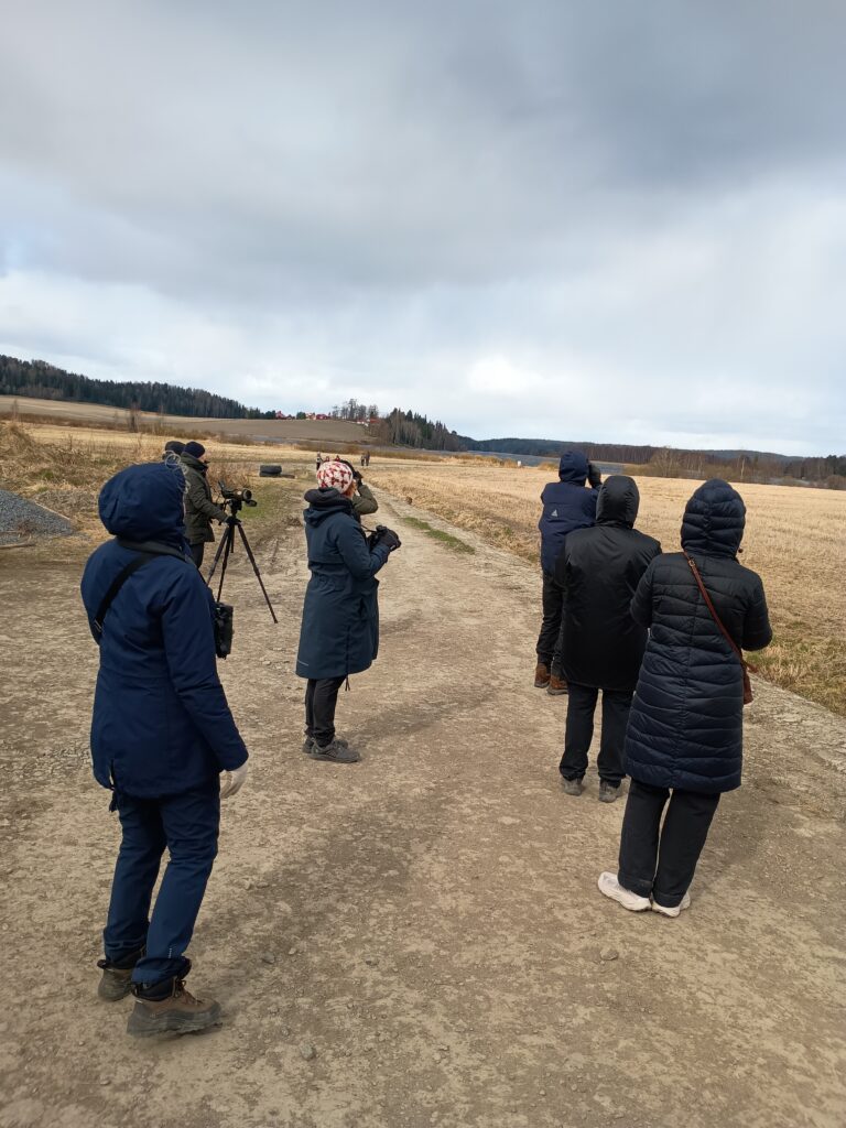 Ryhmä ihmisiä talvisessa ulkoilmassa polulla, osa katselee kiikarilla ja yksi kaukoputkella peltoaukealle.