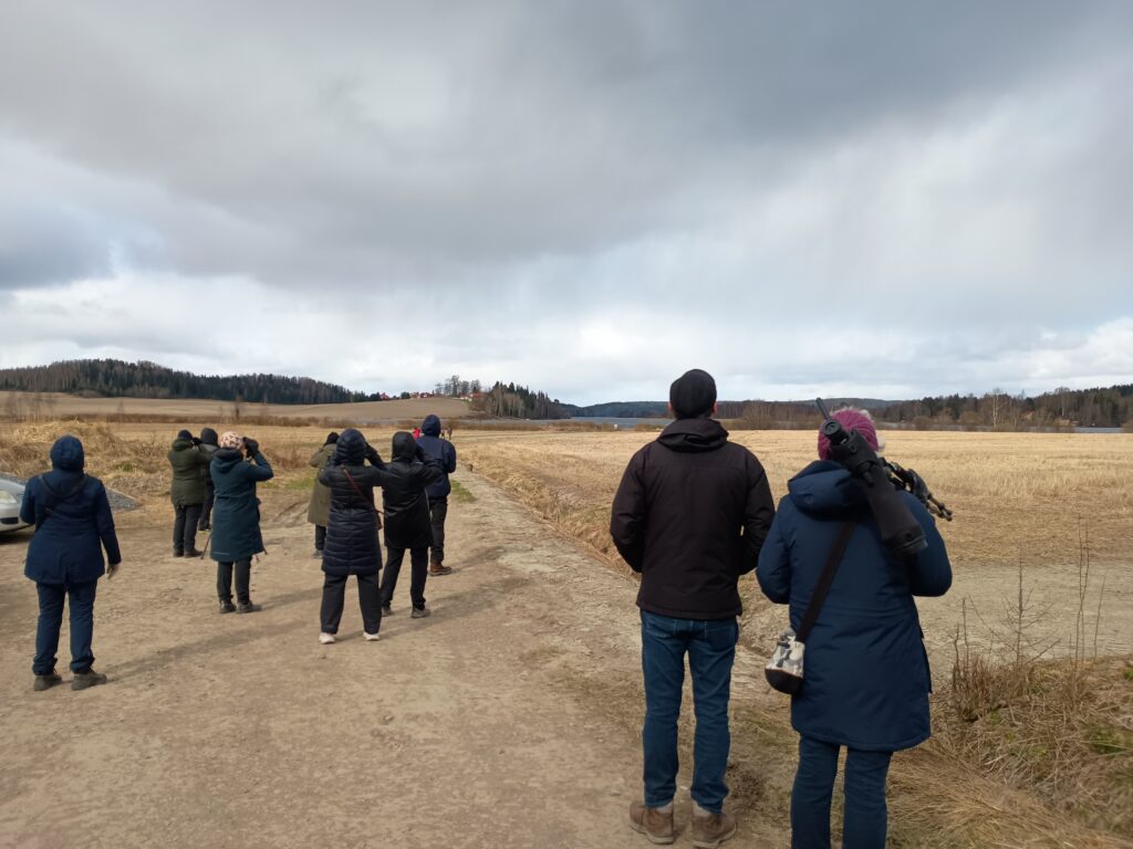 Isompi ryhm&auml; ihmisi&auml; seisoo maatiepolulla ja katsoo kiikarein peltoaukealle, taustalla mets&auml;&auml; ja j&auml;rvimaisema.