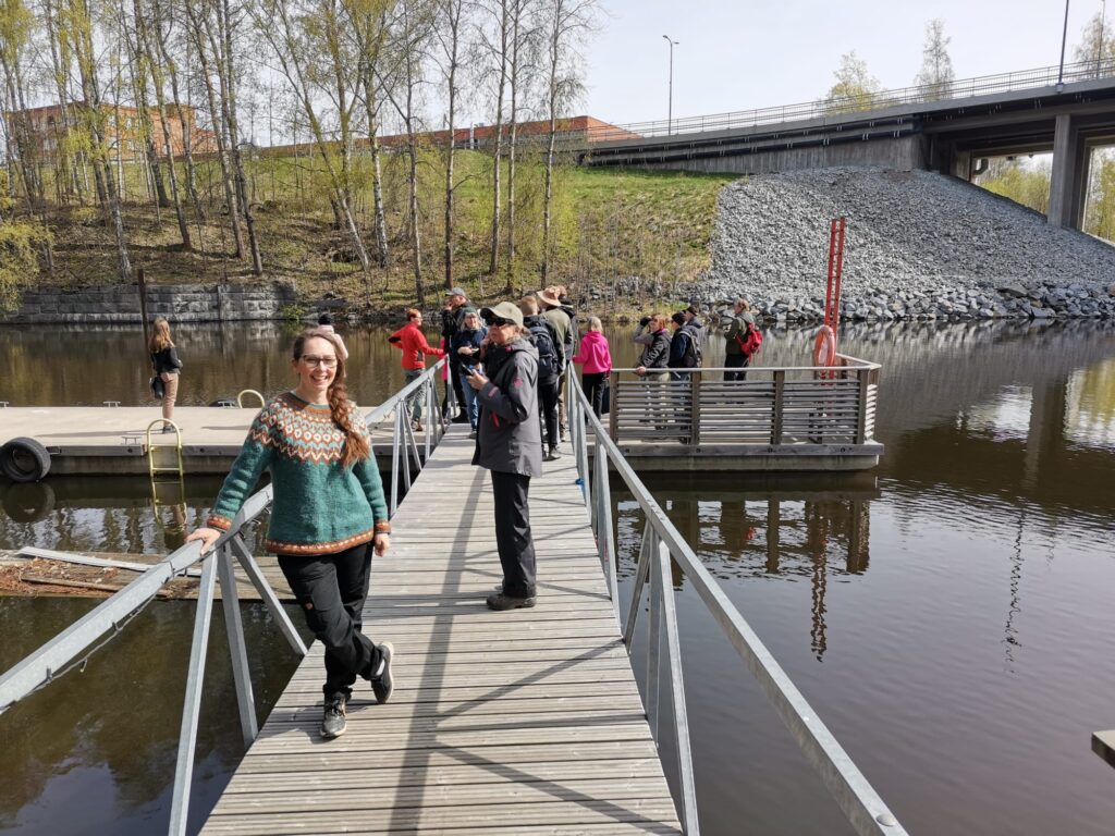 Etualalla seisoo hymyilev&auml; silm&auml;lasip&auml;inen nainen, jolla on p&auml;&auml;ll&auml;&auml;n vihre&auml; kirjoneulepaita ja mustat housut. H&auml;n nojaa k&auml;velysiltaa reunustavaan kaiteeseen. Taustalla n&auml;kyy ryhm&auml; ihmisi&auml; kelluvalla laiturilla sek&auml; harmaakivinen pengerm&auml;, betoninen silta ja kev&auml;isi&auml; koivuja. Taivas on kirkas ja aurinkoinen.