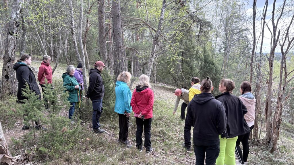 Noin kymmenen hengen ryhmä seisoo metsässä ja kuuntelee opastusta. Taustalla näkyy järvimaisema puiden lomitse.
