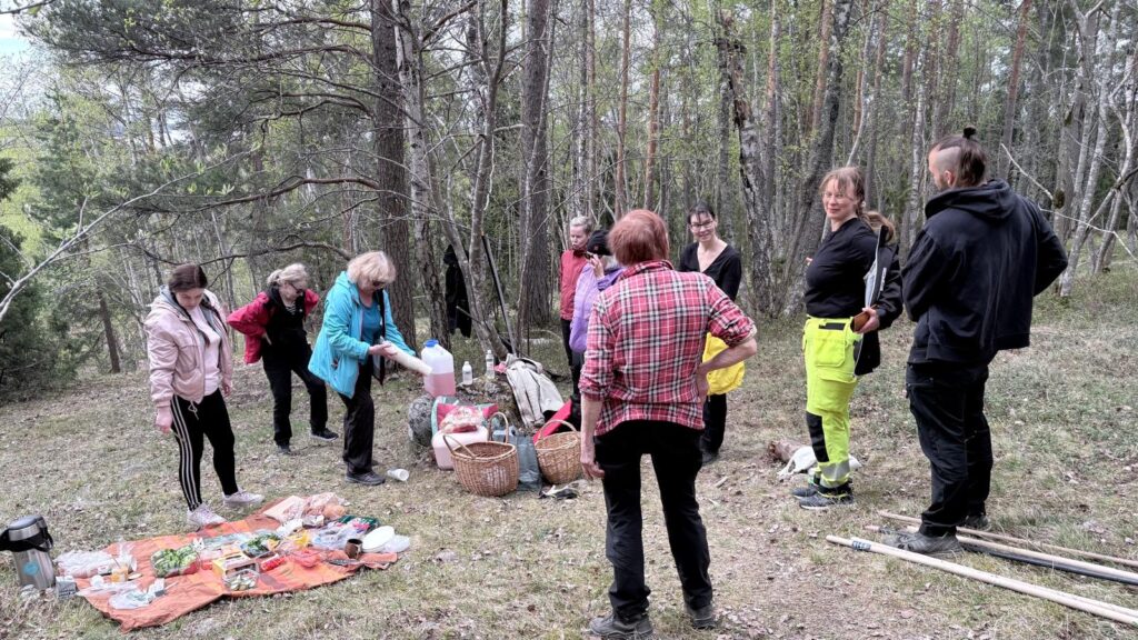 Joukko ihmisiä seisoo metsässä maahan levitetyn eväsdeckin ympärillä. Pöytäliinalla on ruokia ja juomia, ja osallistujat ottavat eväitä.