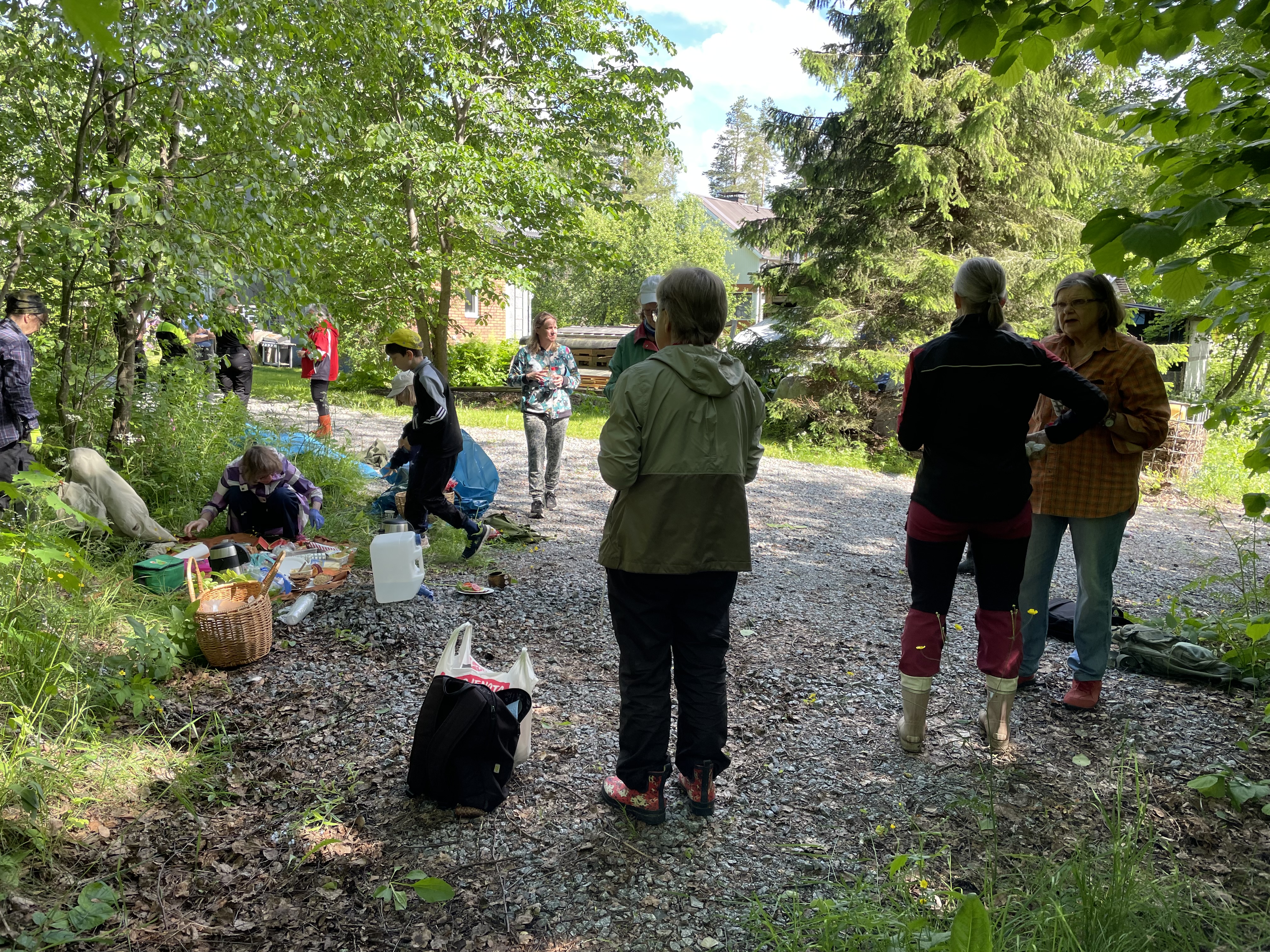Ryhmä ihmisiä kokoontuneena hiekkatielle puiden ja pihapiirin tuntumaan talkoiden yhteydessä. Etualalla muutama aikuinen seisoo keskustelemassa selin kameraan, osalla kumisaappaat. Vasemmalla maassa on piknik-tarvikkeita levitettynä peitteen päälle, ja yksi henkilö istuu kyykyssä niiden äärellä. Keskellä lapsi juoksee ohi keltainen lippalakki päässä. Taustalla näkyy taloja ja havupuita.