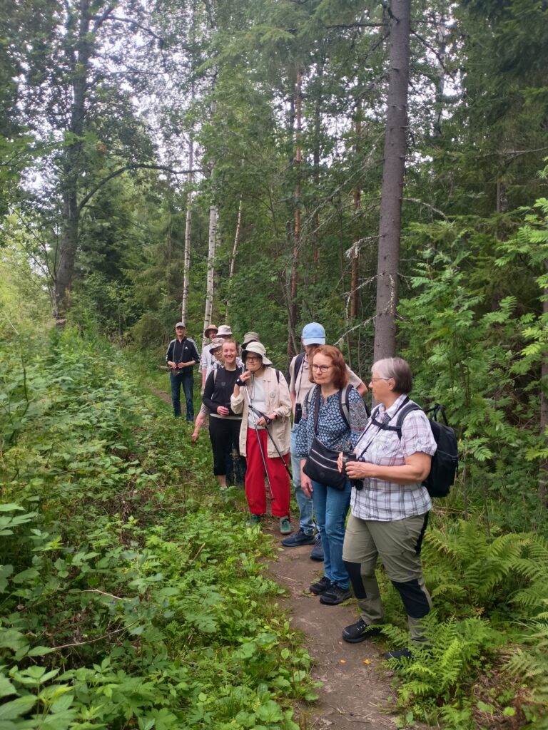 Ryhmä ihmisiä (noin kymmenen) seisoo kapean metsäpolun varrella. Monet lakkeja. Polun molemmin puolin vihreä kasvillisuus.