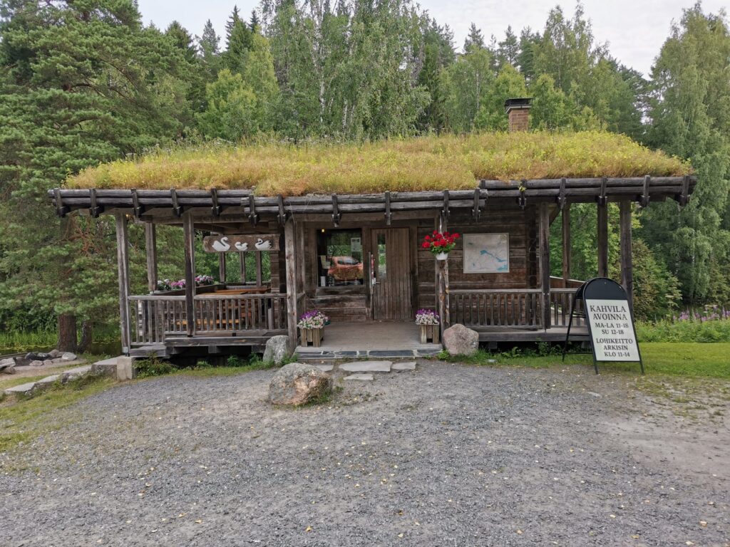Pieni hirsinen kahvila. Katolla kasvaa niittykasveja. Oven edessä kukkalaatikot ja mainoskilpi. Taustalla metsä ja taivas.