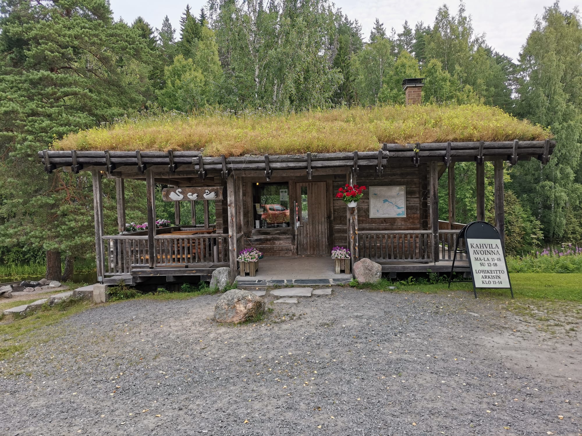 Pieni hirsinen kahvila. Katolla kasvaa niittykasveja. Oven edessä kukkalaatikot ja mainoskilpi. Taustalla metsä ja taivas.