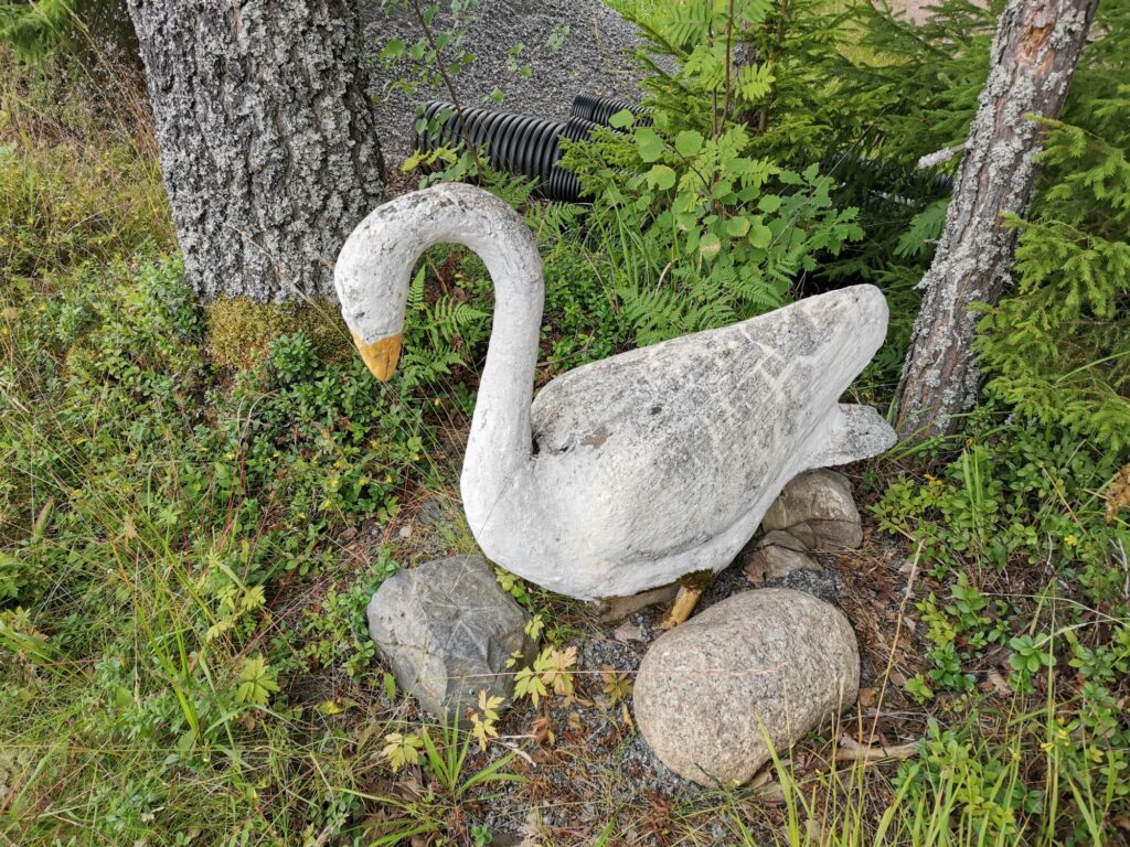 Valkoinen betonista tehty joutsenpatsas kivellä, taustalla metsän kasvillisuutta.