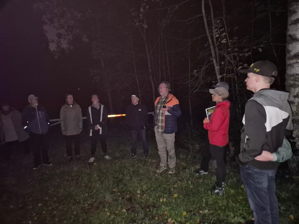 Opas puhuu ryhmälle pimeässä luonnossa, kädessään lepakkodetektori, muut kuuntelevat ympärillä.