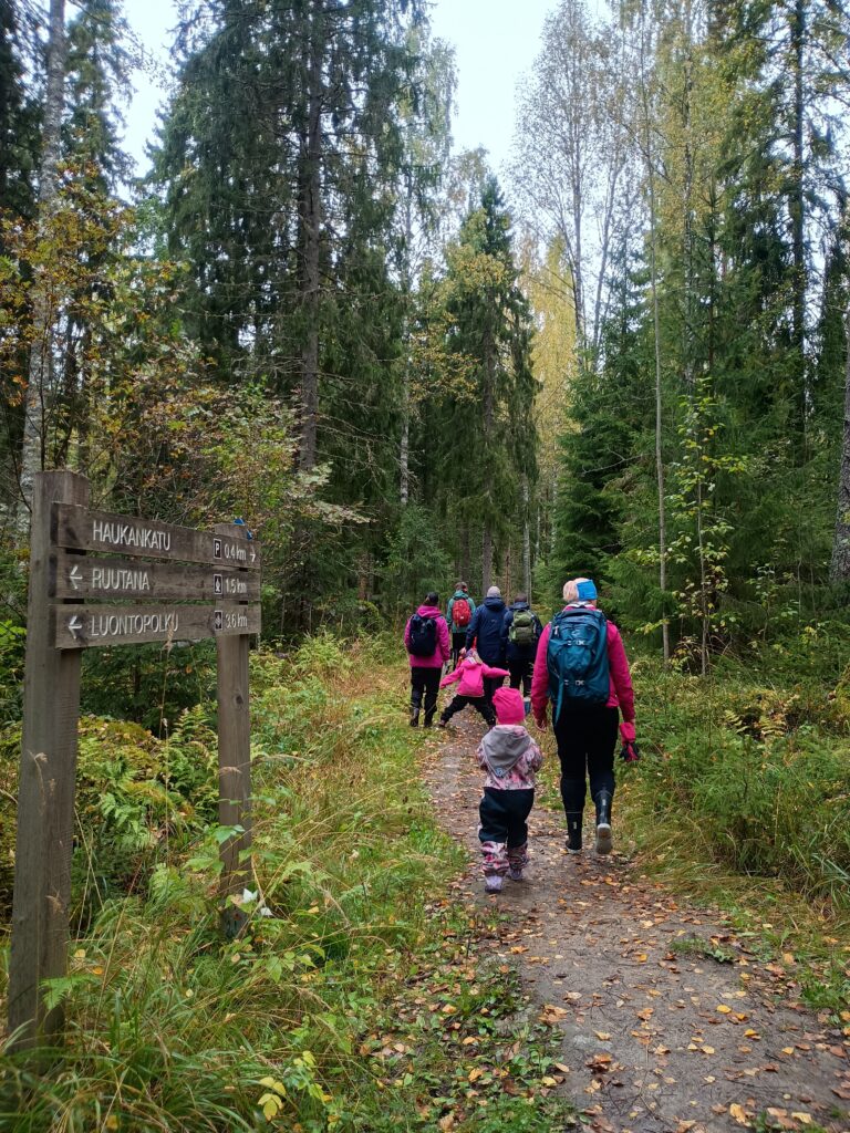 Puinen opastaulu syksyisen mets&auml;polun varressa: Haukankatu, pys&auml;k&ouml;inti 0,4 km oikealle; Ruutana, tulipaikka 1,5 km vasemmalle; Luontopolku 3,6 km vasemmalle. Taustalla ryhm&auml; aikuisia ja lapsia k&auml;velee rinkat sel&auml;ss&auml; pitkin lehtien peitt&auml;m&auml;&auml; hiekkapolkua kuusikkoon p&auml;in.