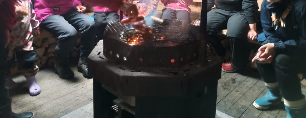 Katetun nuotiopaikan ympärillä istuu lapsia ja aikuisia ulkoiluvaatteissa ja kumisaappaissa. Grillin ritilällä paistuu makkaroita, hiillos hehkuu punaisena ja savu nousee ilmaan.