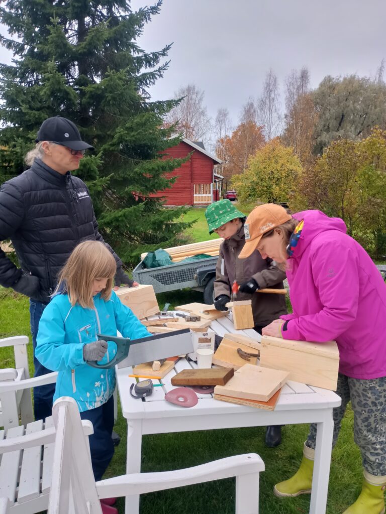 Lapset ja aikuiset rakentavat linnunpönttöjä pihalla valkoisen puutarhapöydän ääressä. Tyttö sahaa lautaa käsisahalla, poika naulaa ja nainen kokoaa pönttöä. Taustalla on punainen hirsitalo ja syksyisiä puita.