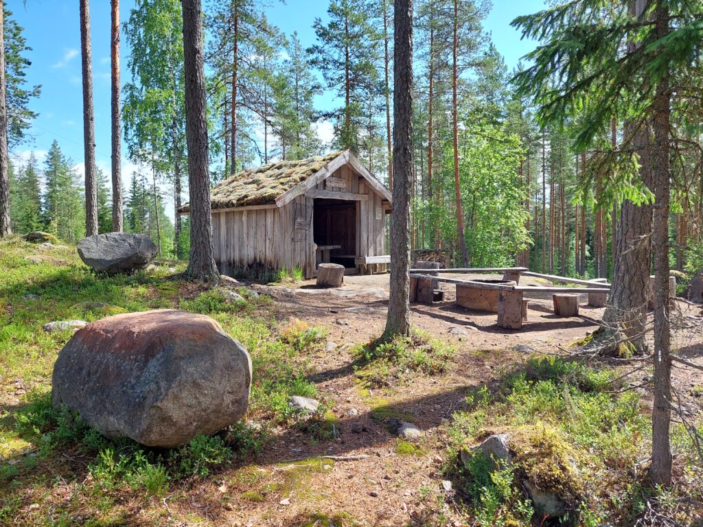 Ramin tölli aurinkoisessa säässä Töllin taipaleen varressa. Töllin edessä on nuotiopaikka.