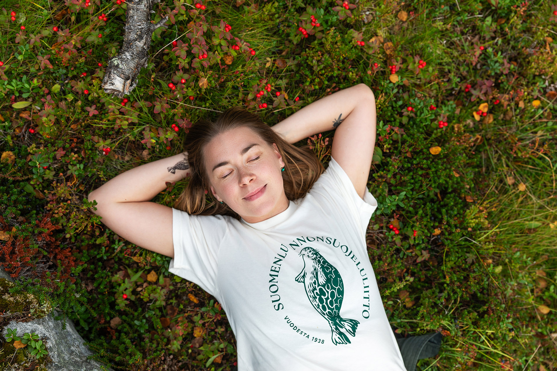 Nainen makaa puolukkamättäällä Luonnonsuojeluliiton paita päällään.