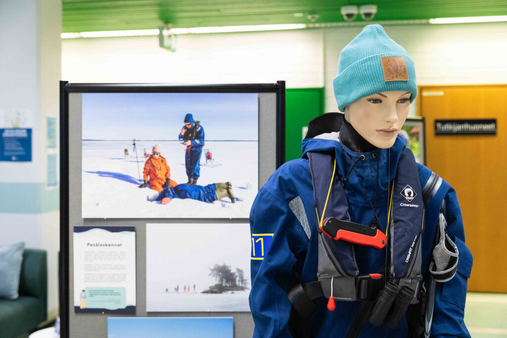 Etualalla mallinukke, jolla on päällään pelastuspuku, naskalit ja paukkuliivit, taustalla valokuvia pesälaskennoista.