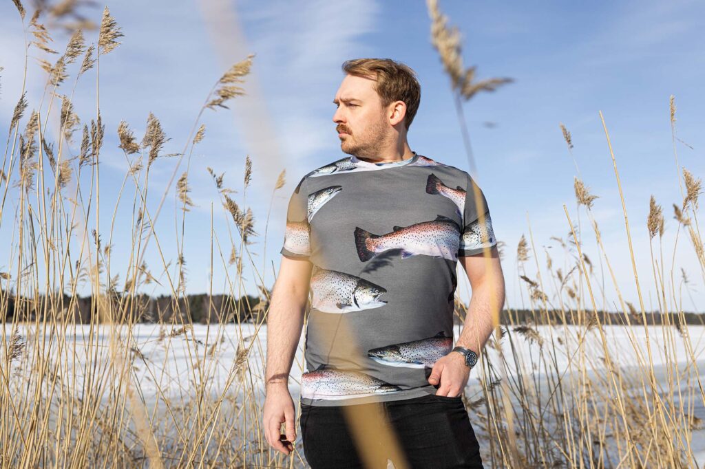 Ihminen seisoo rannalla taimenen kuvilla kuvitetussa T-paidassa.